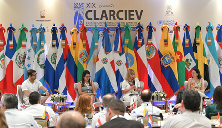 Un panel de discusión con un hombre y tres mujeres. Detrás están las banderas de los países miembros de la OEA y un texto que dice XIX Encuentro de CLARCIEV  