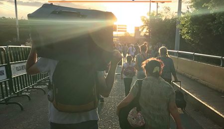 Personas con maletas, bolsos y mochilas se ven de espaldas, a la derecha lo que parece un puente, a la derecha vallas de metal donde se llega a leer "Policía Metropolitana"