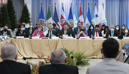 Personas en una mesa. Atrás, las banderas de la OEA, México, Estados Unidos, Costa Rica, Belize, Honduras, Canadá, El Salvador, Guatemala, Panama, la ONU.