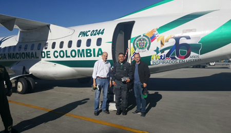 Tres hombres están parados frente a un avión de la Policía Nacional de Colombia, en una pista de aterrizaje. El avión es blanco y verde. Los señores a la izquierda y la derecha están vestidos de civil, mientras que el señor del medio está vestido con